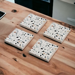 May include: Set of four square coasters with a terrazzo pattern in white, black, gray, and yellow. The coasters are made of a durable material and are perfect for protecting surfaces from heat and scratches.
