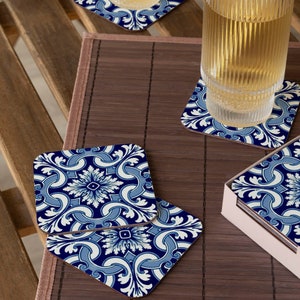May include: Set of blue and white ceramic coasters with a floral pattern. The coasters are square and have a glossy finish. They are arranged on a wooden surface with a glass of liquid.