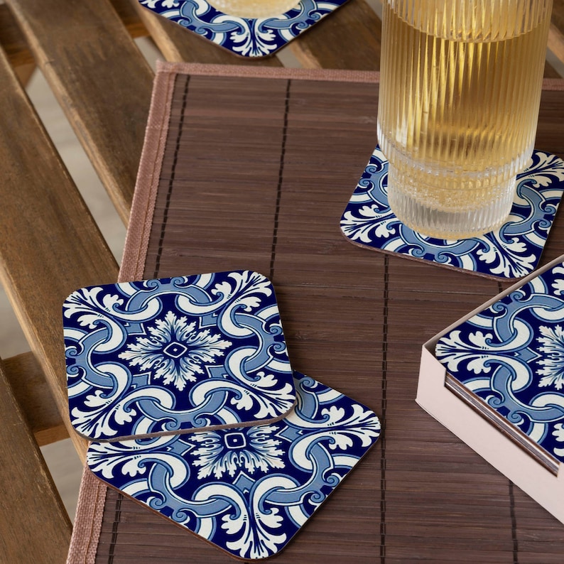 May include: Set of blue and white ceramic coasters with a floral pattern. The coasters are square and have a glossy finish. They are arranged on a wooden surface with a glass of liquid.