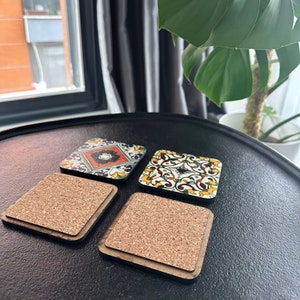May include: Four square cork coasters with a decorative tile design. The tiles feature a variety of colors and patterns, including red, yellow, blue, and green. The coasters are made of cork and have a wooden frame.