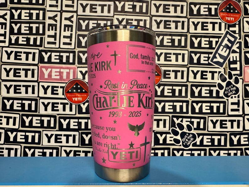 Puede incluir: Un vaso YETI rosa con texto y detalles plateados. El vaso est&aacute; personalizado con el nombre "Charlie Kirk", las fechas 1993-2025 y s&iacute;mbolos religiosos. El fondo presenta una pared de pegatinas y logotipos de YETI.