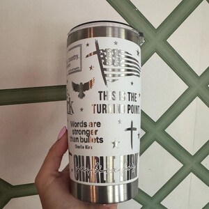 Puede incluir: Un vaso de acero inoxidable con una envoltura blanca con un dise&ntilde;o de bandera estadounidense, cruz y &aacute;guila. Tambi&eacute;n se muestra el texto "THIS IS THE TURNING POINT" y "Words are stronger than bullets". El vaso est&aacute; sostenido por una persona.