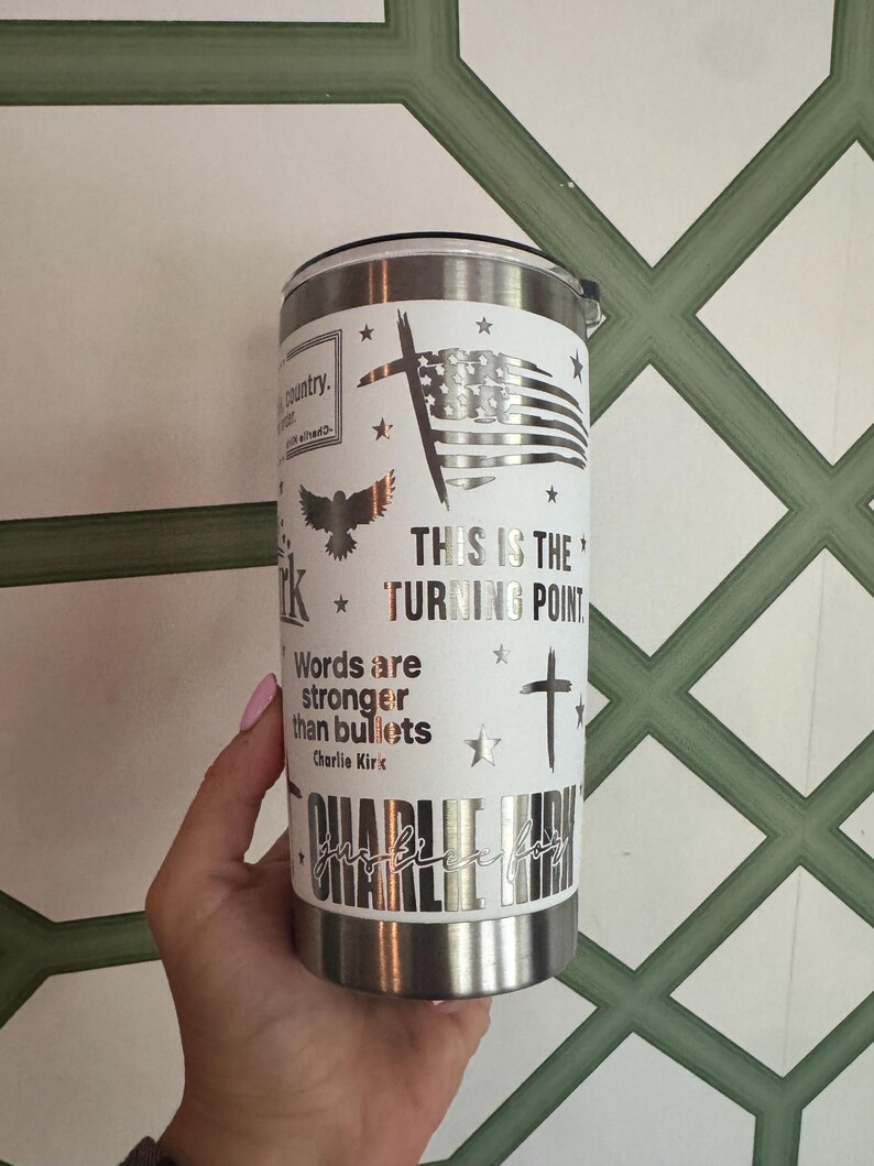 Puede incluir: Un vaso de acero inoxidable con una envoltura blanca con un dise&ntilde;o de bandera estadounidense, cruz y &aacute;guila. Tambi&eacute;n se muestra el texto "THIS IS THE TURNING POINT" y "Words are stronger than bullets". El vaso est&aacute; sostenido por una persona.