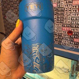 May include: A blue Yeti tumbler with a gold logo and a white wave design. The tumbler is being engraved with a laser.