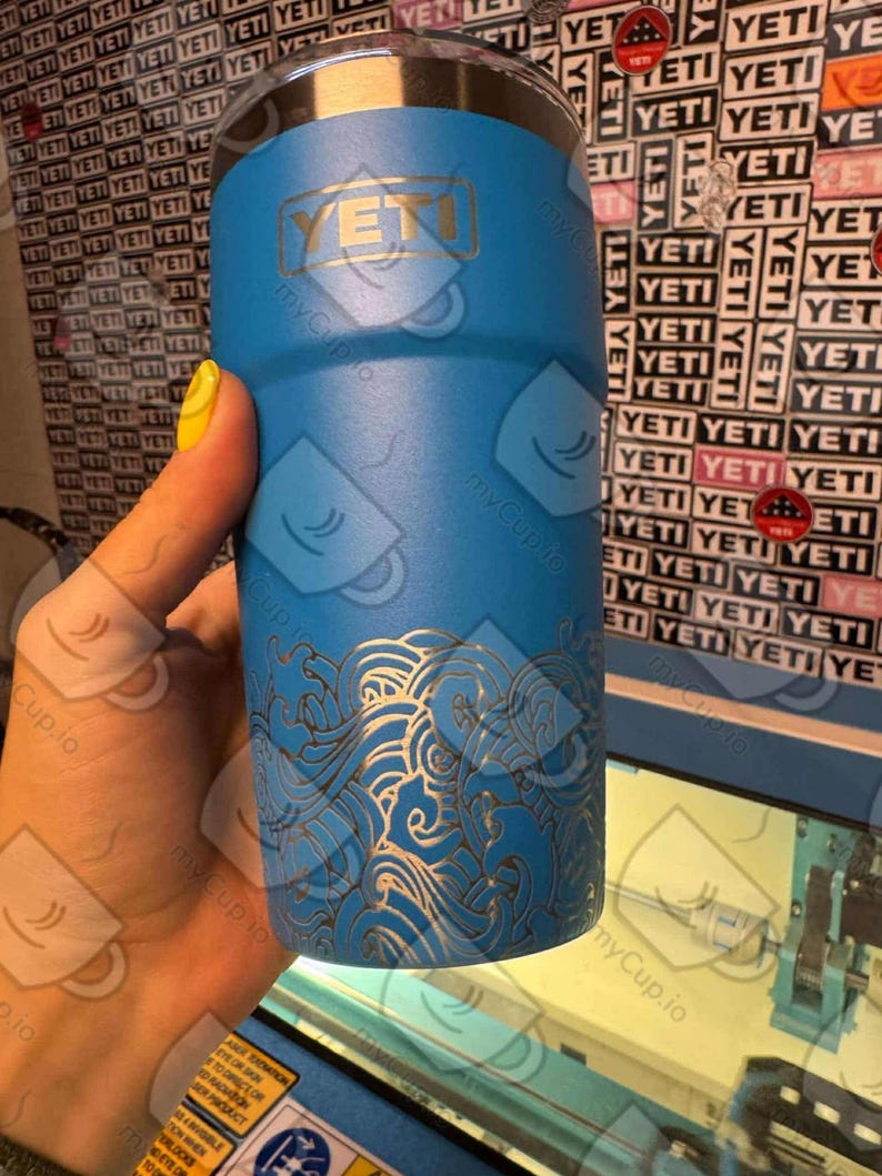 May include: A blue Yeti tumbler with a gold logo and a white wave design. The tumbler is being engraved with a laser.