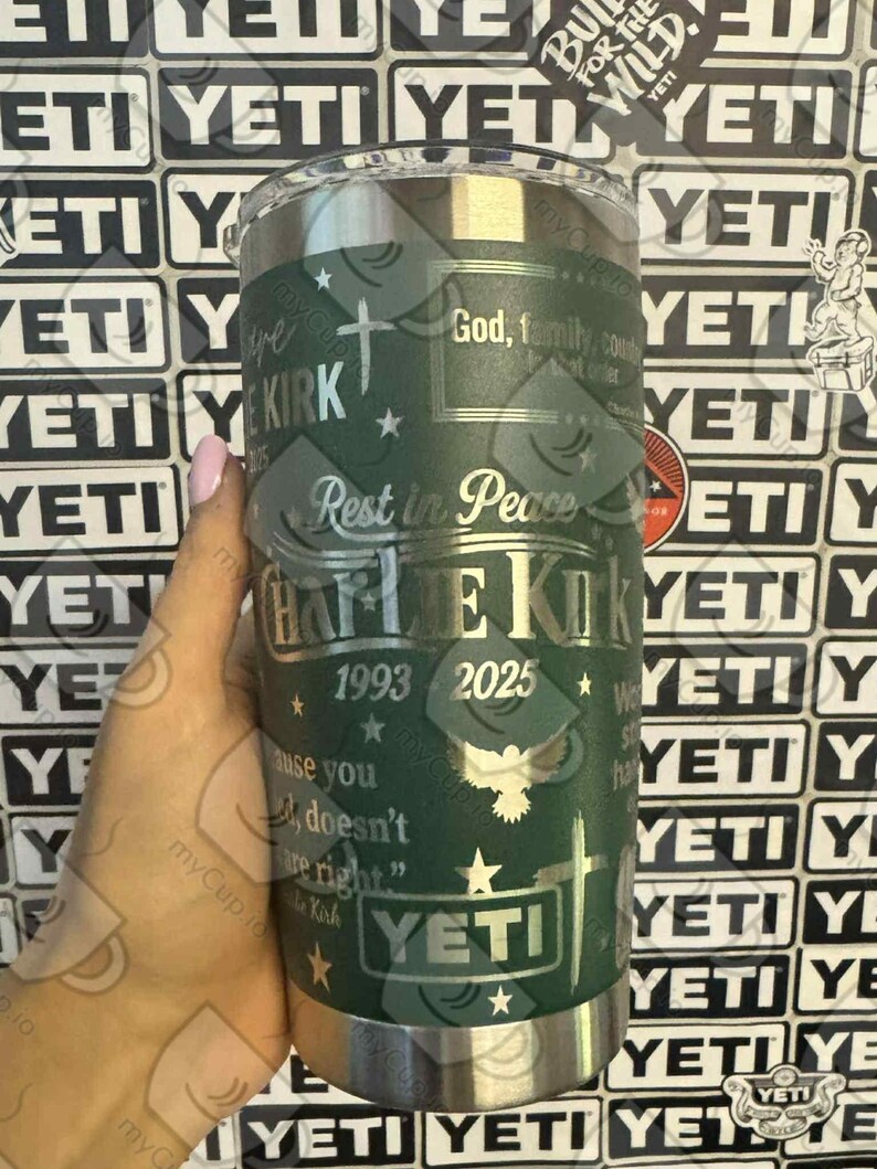 Puede incluir: Un vaso YETI verde con texto y detalles plateados. El texto incluye "Charlie Kirk", "1993-2025", una cruz, estrellas y las palabras "Rest in Peace". El vaso tiene un borde plateado y una tapa transparente.