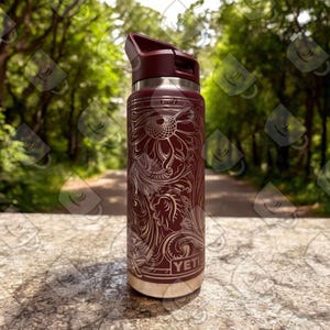 Puede incluir: Una YETI Rambler de color marr&oacute;n con un dise&ntilde;o floral grabado en acero inoxidable. La botella tiene una tapa negra con un asa de bucle.