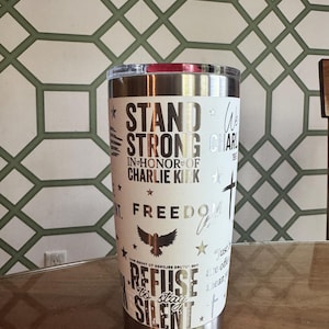 Puede incluir: Un vaso de acero inoxidable blanco con tapa plateada. El vaso presenta las palabras "STAND STRONG" y "REFUSE TO STAY SILENT" en letras plateadas, junto con otros s&iacute;mbolos patri&oacute;ticos. El vaso es cil&iacute;ndrico y est&aacute; sobre una superficie de madera.