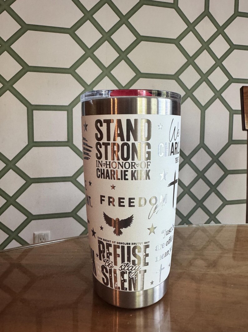 Puede incluir: Un vaso de acero inoxidable blanco con tapa plateada. El vaso presenta las palabras "STAND STRONG" y "REFUSE TO STAY SILENT" en letras plateadas, junto con otros s&iacute;mbolos patri&oacute;ticos. El vaso es cil&iacute;ndrico y est&aacute; sobre una superficie de madera.