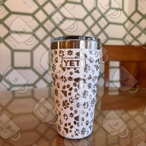 Puede incluir: Un vaso YETI blanco con una tapa plateada y un diseño de huellas de patas. El vaso está sobre una mesa de madera.