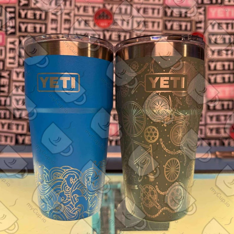 May include: Two YETI brand stainless steel tumblers with lids. The tumbler on the left is blue with a gold wave design. The tumbler on the right is green with a gold bicycle design.