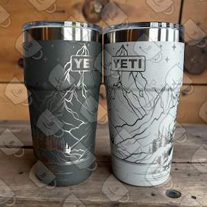 Pode incluir: Dois copos Yeti de aço inoxidável com desenhos de montanhas e florestas. Um é cinza escuro, o outro cinza claro. Cada copo tem uma borda prateada e o logotipo Yeti.