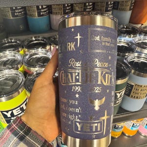 Puede incluir: Un vaso YETI de acero inoxidable con un acabado texturizado azul marino. El vaso presenta texto y gr&aacute;ficos plateados, incluyendo una cruz, una paloma y el nombre "Charlie Kirk". El texto incluye las frases "Dios, familia, en ese orden" y "Descansa en paz."