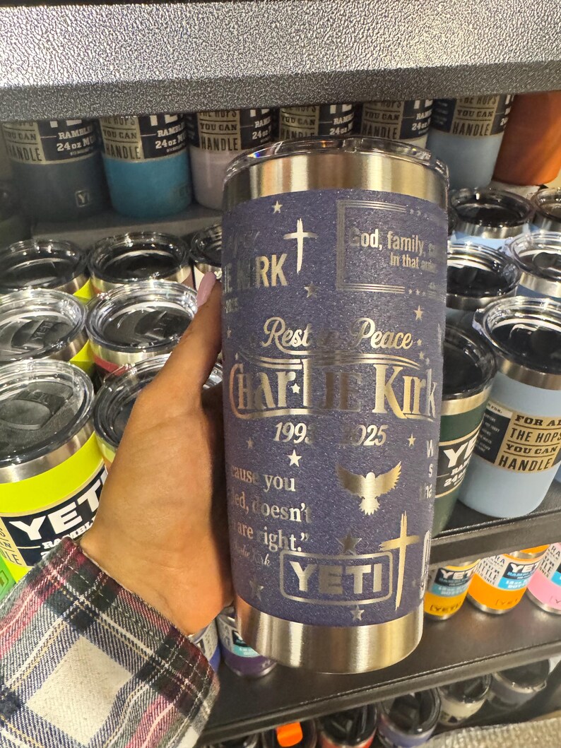 Puede incluir: Un vaso YETI de acero inoxidable con un acabado texturizado azul marino. El vaso presenta texto y gr&aacute;ficos plateados, incluyendo una cruz, una paloma y el nombre "Charlie Kirk". El texto incluye las frases "Dios, familia, en ese orden" y "Descansa en paz."