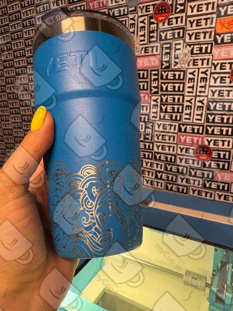 May include: A blue Yeti tumbler with a laser-etched design of waves. The tumbler has a silver lid and the Yeti logo is visible on the side.