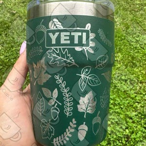 Puede incluir: Un vaso YETI verde con tapa transparente, con un diseño de hojas y bellotas plateadas. El vaso se sostiene sobre un fondo de hierba verde. El logotipo de YETI es visible.