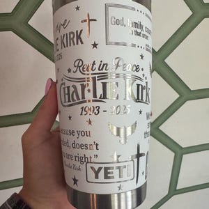 Puede incluir: Un vaso Yeti de acero inoxidable con una envoltura blanca con el texto "Rest in Peace Charlie Kirk 1993-2025", cruces, estrellas y un gr&aacute;fico de &aacute;guila. El vaso est&aacute; sostenido por una persona.
