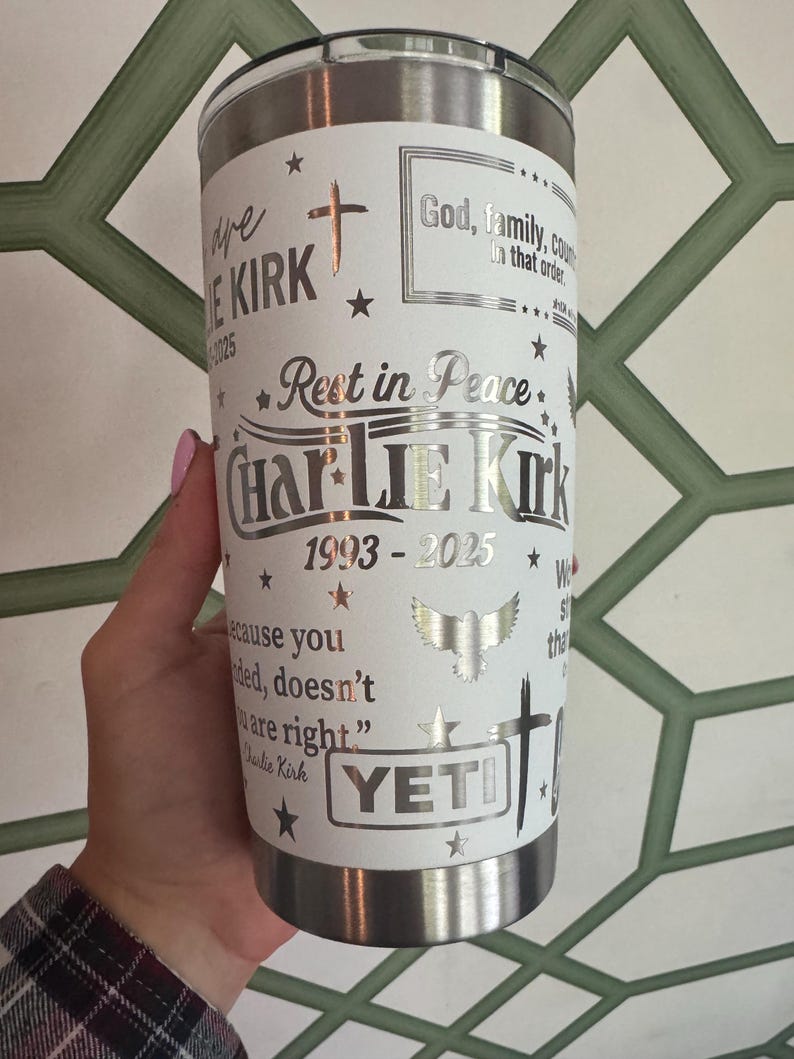 Puede incluir: Un vaso Yeti de acero inoxidable con una envoltura blanca con el texto "Rest in Peace Charlie Kirk 1993-2025", cruces, estrellas y un gr&aacute;fico de &aacute;guila. El vaso est&aacute; sostenido por una persona.
