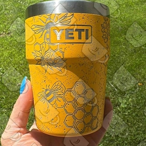 Op de afbeelding: Gele YETI-beker met een roestvrijstalen rand, met gegraveerde bijen- en honingraatontwerpen. De beker heeft het YETI-logo. De achtergrond is groen gras. De beker wordt vastgehouden in een hand met een blauw geschilderde nagel.