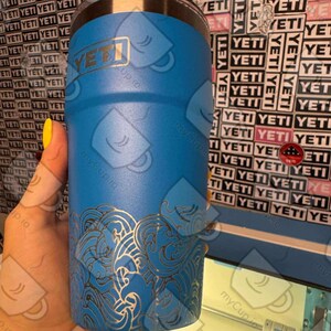 May include: A blue Yeti tumbler with a laser-etched design of waves. The tumbler has a stainless steel lid and a Yeti logo on the side.