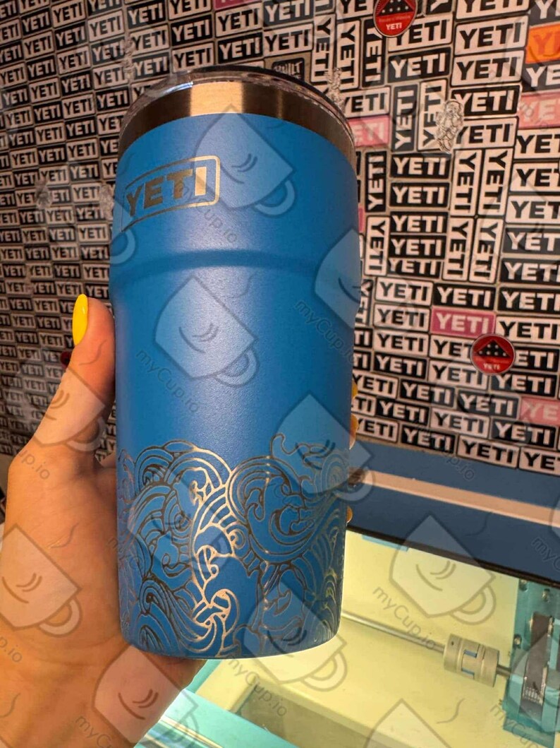 May include: A blue Yeti tumbler with a laser-etched design of waves. The tumbler has a stainless steel lid and a Yeti logo on the side.