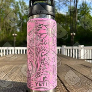 Puede incluir: YETI Rambler rosa con un dise&ntilde;o floral dorado. La botella tiene una tapa negra y una asa de transporte.