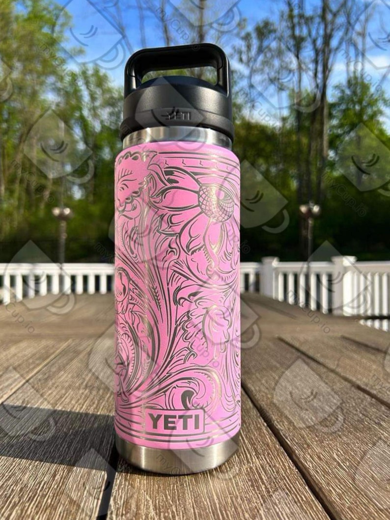 Puede incluir: YETI Rambler rosa con un dise&ntilde;o floral dorado. La botella tiene una tapa negra y una asa de transporte.
