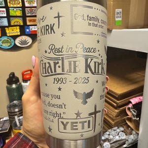 Puede incluir: Un vaso YETI beige con texto y dise&ntilde;os grabados. El texto incluye "Rest in Peace", "Charlie Kirk 1993-2025" y otras frases. Presenta una cruz, estrellas y un p&aacute;jaro volando. El vaso es sostenido por una persona.
