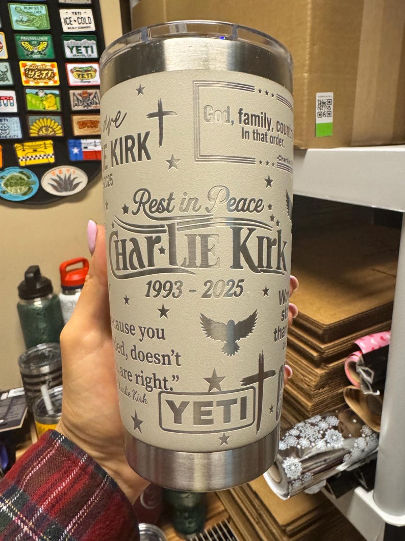 Puede incluir: Un vaso YETI beige con texto y dise&ntilde;os grabados. El texto incluye "Rest in Peace", "Charlie Kirk 1993-2025" y otras frases. Presenta una cruz, estrellas y un p&aacute;jaro volando. El vaso es sostenido por una persona.