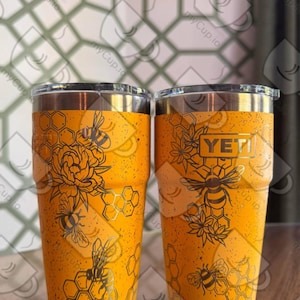 Op de afbeelding: Twee gele geïsoleerde bekers met zwarte bijen- en honingraatontwerpen. Eén beker heeft een bloemenontwerp met bijen, de andere toont het "YETI"-logo en bijengraphics. Beide hebben zilveren randen en deksels.