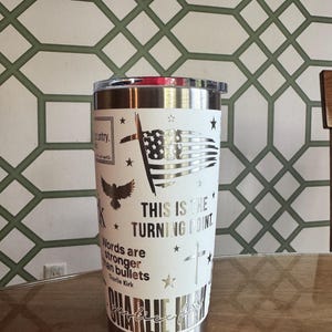 Puede incluir: Un vaso de acero inoxidable con una envoltura blanca con una bandera estadounidense, una cruz y texto. El texto incluye "THIS IS THE TURNING POINT" y una cita de Charlie Kirk. El vaso tiene un borde y una tapa plateados.