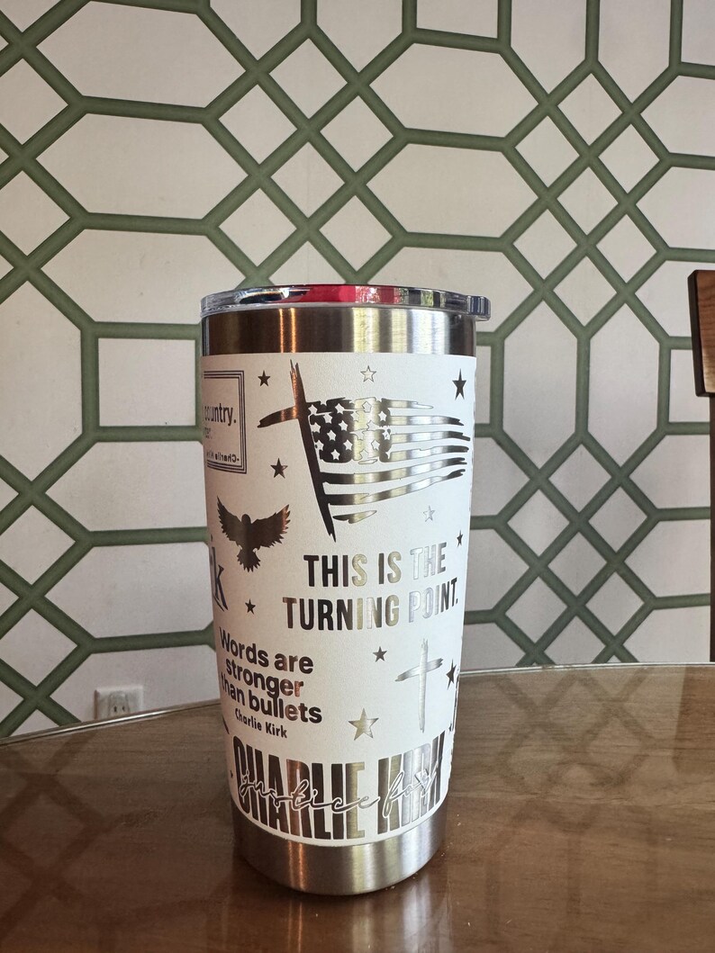 Puede incluir: Un vaso de acero inoxidable con una envoltura blanca con una bandera estadounidense, una cruz y texto. El texto incluye "THIS IS THE TURNING POINT" y una cita de Charlie Kirk. El vaso tiene un borde y una tapa plateados.