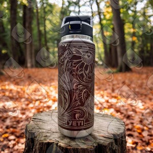 Puede incluir: Una botella de agua YETI de acero inoxidable con un dise&ntilde;o floral marr&oacute;n y blanco. La botella est&aacute; sentada sobre un toc&oacute;n de &aacute;rbol en un entorno boscoso.