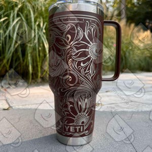 May include: A brown Yeti tumbler with a silver floral design and a black handle. The tumbler has a straw lid and the Yeti logo is visible on the front.