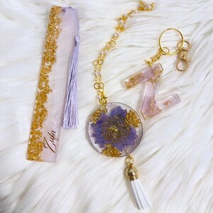 May include: A collection of handmade resin accessories, including a bookmark, a keychain with a floral design, and a letter "Z" keychain. The items feature gold leaf accents and a light purple hue. The bookmark has a tassel and the name "Zufa" is written on it.