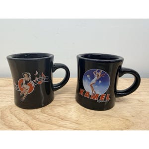May include: Two black ceramic mugs with handles. The left mug features a pin-up style illustration of a person dancing with musical notes. The right mug displays a circular graphic of a person skydiving with the text KAMEL Cigarettes printed below.