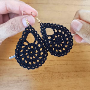 Puede incluir: Un par de pendientes de ganchillo negros en forma de lágrima con ganchos dorados. Los pendientes presentan un diseño calado detallado con un patrón en espiral en el centro. Los pendientes son hechos a mano.