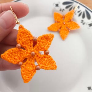 May include: A pair of handmade orange crochet flower earrings with gold-colored hooks. Each earring features a five-petal flower design with small, copper-colored beads at the center of each petal. The earrings are displayed on a white plate.