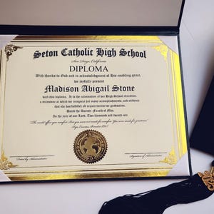 May include: A diploma from Seton Catholic High School, featuring gold accents and the name Madison Abigail Stone. The diploma is in a black case, with a graduation cap and tassel visible.