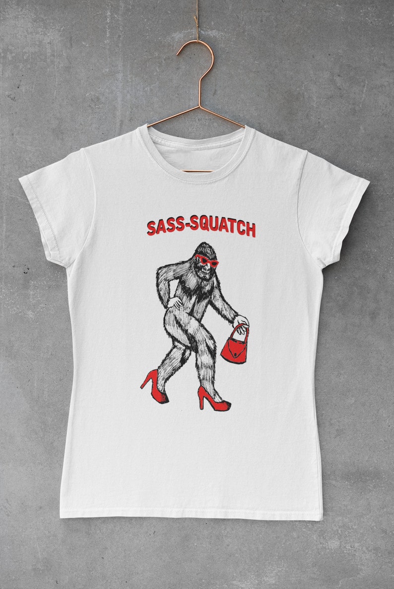 Puede incluir: Camiseta blanca con el texto "SASS-SQUATCH" en rojo. El gr&aacute;fico presenta una criatura parecida a Bigfoot con tacones rojos, gafas de sol y un bolso rojo. La camiseta est&aacute; colgada de una percha de color cobre.
