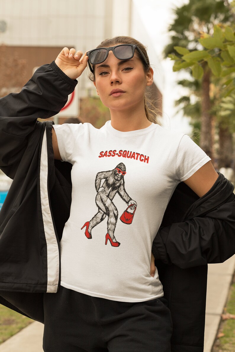Puede incluir: Camiseta blanca con el texto "Sass-Squatch" en rojo. La camiseta presenta un gr&aacute;fico de una criatura parecida a Bigfoot con tacones altos y gafas de sol rojos, llevando un bolso rojo. La camiseta es un dise&ntilde;o humor&iacute;stico.