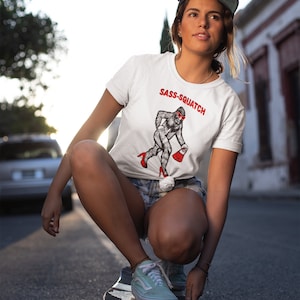 Puede incluir: Camiseta blanca con el texto "Sass-Squatch" y un dibujo de una criatura parecida a Pie Grande con gafas de sol y tacones rojos, que lleva un bolso rojo. La persona lleva la camiseta en un monopat&iacute;n.