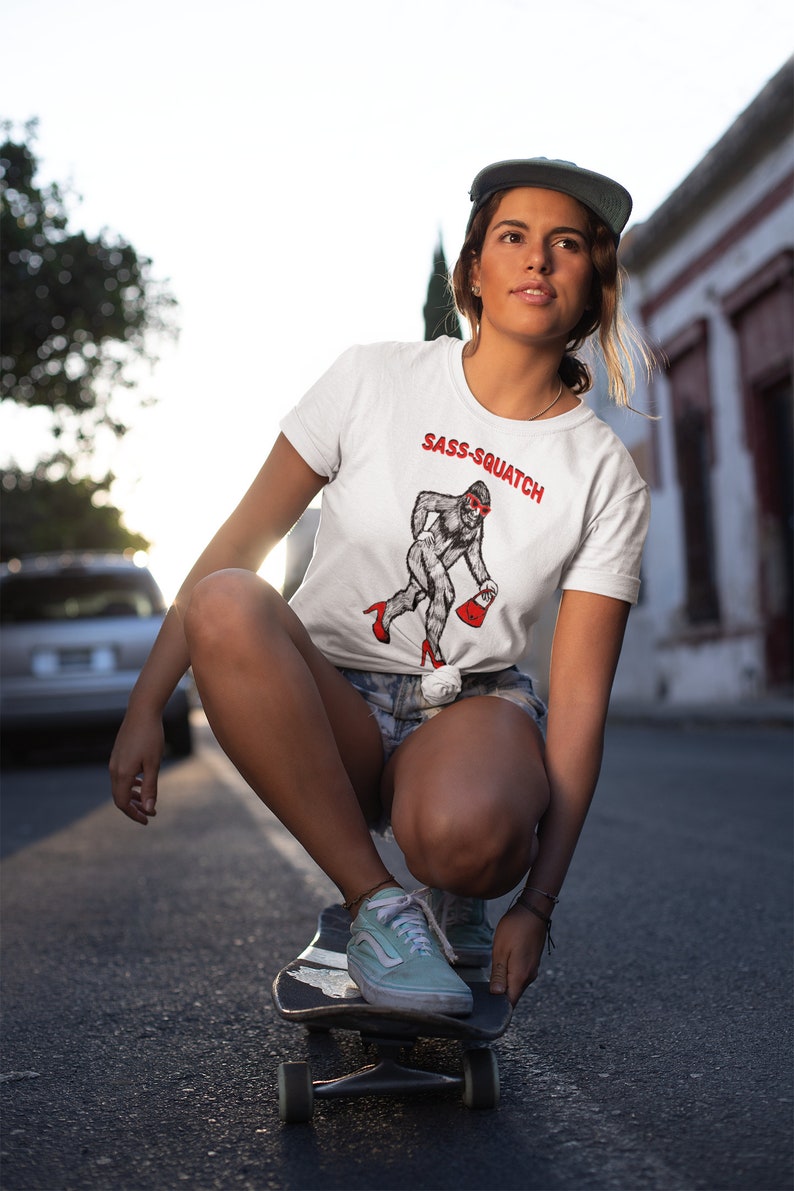 Puede incluir: Camiseta blanca con el texto "Sass-Squatch" y un dibujo de una criatura parecida a Pie Grande con gafas de sol y tacones rojos, que lleva un bolso rojo. La persona lleva la camiseta en un monopat&iacute;n.