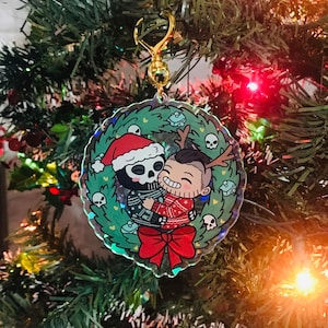Pode incluir: Um enfeite de Natal redondo e transparente com um fecho dourado, pendurado numa árvore de Natal verde. O enfeite apresenta uma ilustração de desenho animado de duas figuras abraçadas, rodeadas por uma coroa de caveiras e corações e um laço vermelho.