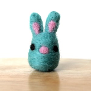 May include: A small, teal blue felt bunny with pink ears and a pink nose. The bunny has black eyes and is sitting on a wooden surface.