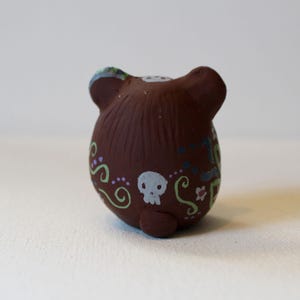 K&ouml;nnte beinhalten: Eine kleine, runde Tonfigur in Form eines B&auml;ren mit abgerundeten Ohren. Die Oberfl&auml;che ist mit handgemalten Mustern verziert, darunter ein wei&szlig;er Totenkopf, hellgr&uuml;ne Schn&ouml;rkel, violette Punkte und kleine florale Verzierungen am K&ouml;rper.