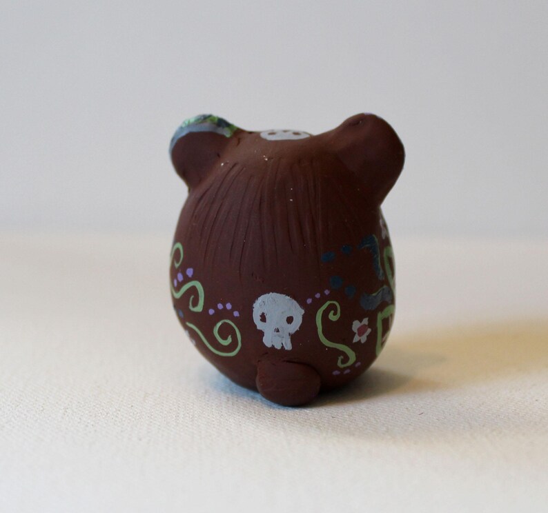 K&ouml;nnte beinhalten: Eine kleine, runde Tonfigur in Form eines B&auml;ren mit abgerundeten Ohren. Die Oberfl&auml;che ist mit handgemalten Mustern verziert, darunter ein wei&szlig;er Totenkopf, hellgr&uuml;ne Schn&ouml;rkel, violette Punkte und kleine florale Verzierungen am K&ouml;rper.