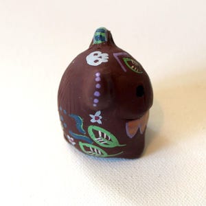 K&ouml;nnte beinhalten: Eine kleine, braune, abgerundete Tonfigur mit handgemalten Details, darunter ein wei&szlig;er Totenkopf, gr&uuml;ne Bl&auml;tter, kleine Blumen und violette Punkte. Das Objekt hat eine glatte, matte Textur und eine kleine Schlaufe an der Oberseite.