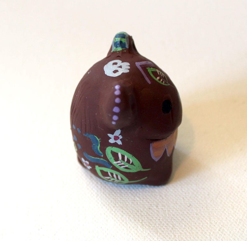 K&ouml;nnte beinhalten: Eine kleine, braune, abgerundete Tonfigur mit handgemalten Details, darunter ein wei&szlig;er Totenkopf, gr&uuml;ne Bl&auml;tter, kleine Blumen und violette Punkte. Das Objekt hat eine glatte, matte Textur und eine kleine Schlaufe an der Oberseite.