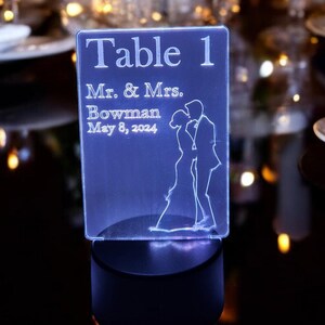 May include: A clear acrylic table number with a white LED light base. The number is "Table 1" and the text reads "Mr. & Mrs. Bowman May 8, 2024". A silhouette of a couple is shown on the right side of the sign.